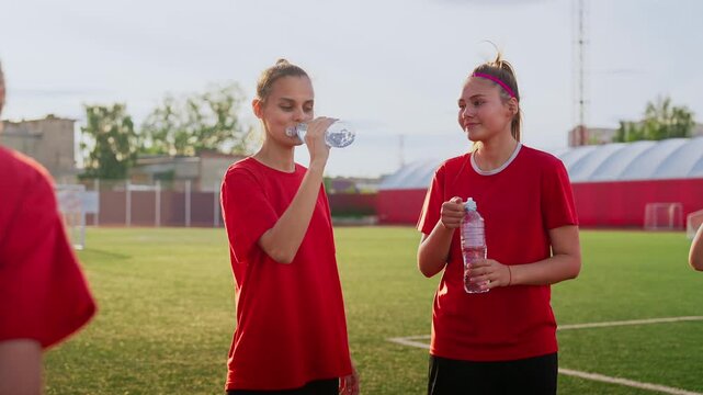 Female soccer players in red jerseys hydrate and recover together on a sunny outdoor field during team training break, sharing smiles, teamwork, and athletic energy