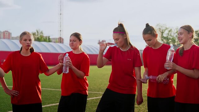 Womens soccer team taking a hydration break during outdoor training session on sunny field, teammates in red jerseys resting and drinking water together