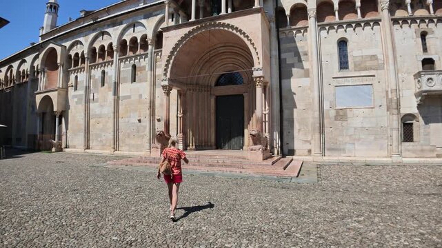 Woman with two pigtails standing with her back to the viewer. Looking at the elaborate Romanesque architecture and detailed carvings of Modena Cathedral under a clear blue sky of Modena city in Italy