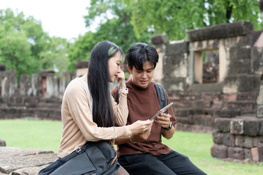 Young Asian travelers using digital tablet while exploring ancient stone ruins, planning their journey, discovering cultural heritage, enjoying outdoor adventure, and smart travel experiences.