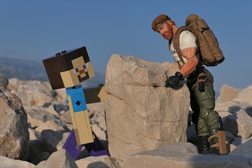 Obraz premium Action figures of GI JOE Classified edtition survival instructor Outback helping Minecraft hero Steve to place a large square footstone on Croatian beach, summer daylight sunshine. 