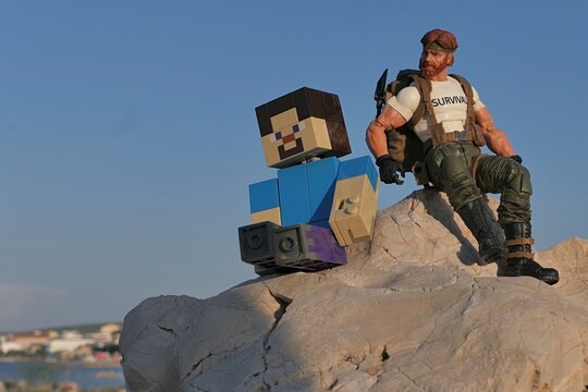 Action figures of Minecraft hero Steve and GI JOE Classified series survival instructor Outback sitting on coastal rock in Croatia, sunlit by evening sunshine, blue skies in background. 