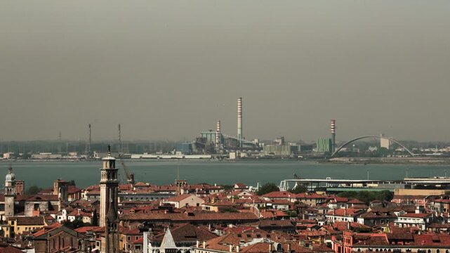 Historic terracotta rooftops overlook the calm lagoon towards tall industrial factory smokestacks. A striking visual contrast inspiring corporate sustainability and environmental awareness.