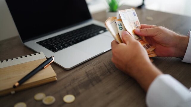 Unrecognizable businessman counting a stack of euro banknotes at an office desk with a laptop and notebook, representing salary, savings, income, personal finance and financial success.
