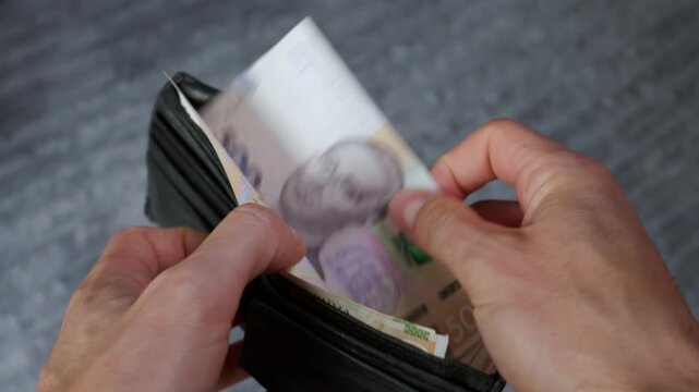 A close-up of male hands opening a black leather wallet filled with Ukrainian hryvnia banknotes. The person is counting their paper money against a grey textured background.