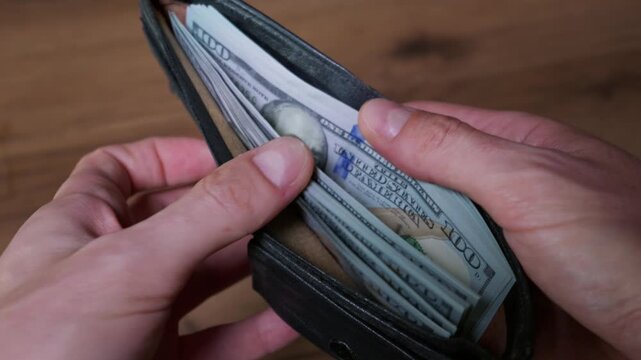 Hands open a black leather wallet packed with a thick stack of United States hundred-dollar bills. Benjamin Franklin portrait is visible on the currency over a wooden table background.