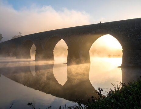 Ancient stone bridge at sunrise with morning mist, historic arched bridge reflecting in calm river water, atmospheric foggy landscape at dawn