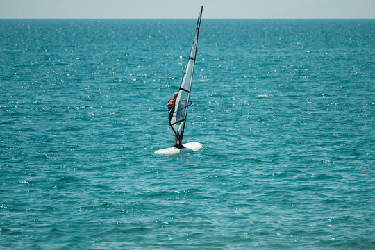 a lone windsurfer riding a white board with a tall sail across a vast, sparkling blue ocean under a clear sky.