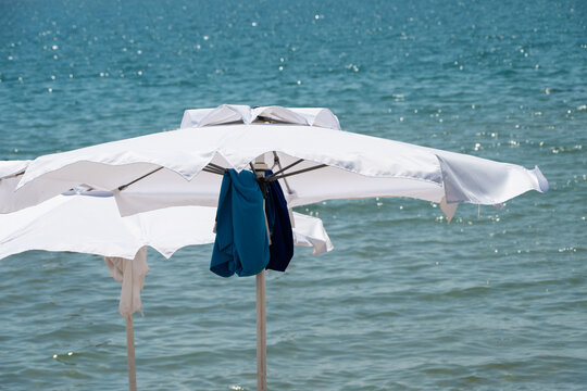 bright white beach umbrellas providing shade, with a dark blue towel hanging casually from the metal frame against the backdrop of a sparkling, sunlit ocean.