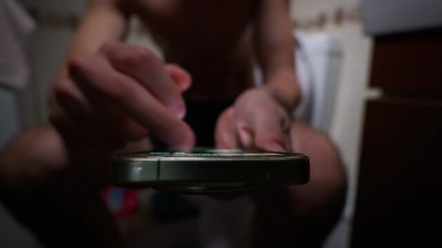 A shirtless man sits on a toilet in a dimly lit bathroom while his finger scrolls through social media content on a glowing smartphone screen. Concept of digital addiction, doomscrolling, and habit