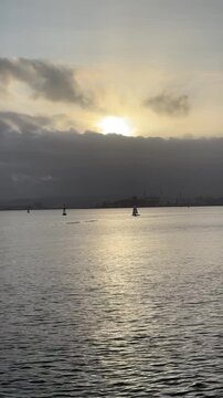 A small sailboat glides across calm coastal waters as golden sunset reflections contrast with layered gray clouds, revealing the unique light of a peaceful evening at sea.