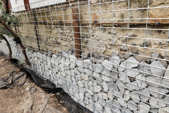 Stone fence in steel mesh and green lawn. Close up of gabion with tightly packed stones of varying sizes and textures. Combination of natural materials and industrial design.