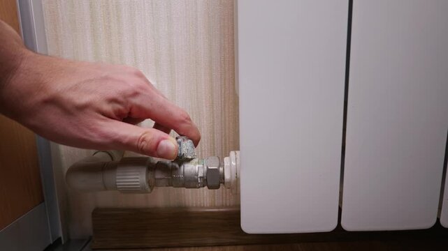 A man hand reaches out to turn a small metal valve connected to a white wall-mounted radiator. The plumbing fixtures and pipes are visible against a beige wallpaper background in the room.