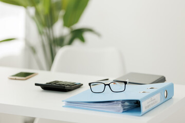 Minimal bright office workspace featuring a blue binder with documents, reading glasses, calculator...