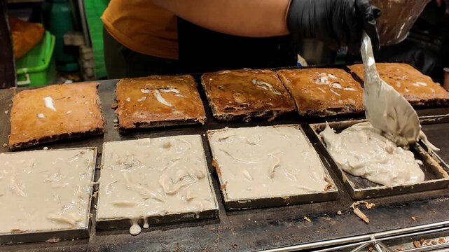 Close-up of a street food vendor spreading coconut batter inside a square mold while preparing a traditional Thai coconut dessert at a busy night market in Phuket, Thailand. Authentic Asian street foo