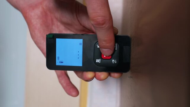 A hand holds a black digital laser distance measure tool flat against a smooth wooden surface. The blue illuminated LCD screen displays a reading of zero point zero zero zero meters.