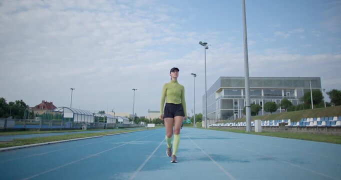 A young beautiful girl in sportswear and a cap with a skipping rope in her hands walks along the stadium's running track to train and exercise on a summer day