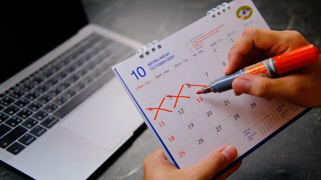 Vacation planning calendar with a laptop on the office desk while a hand marks off days using a red marker. Represents annual leave planning, long holiday scheduling, paid time off (PTO).