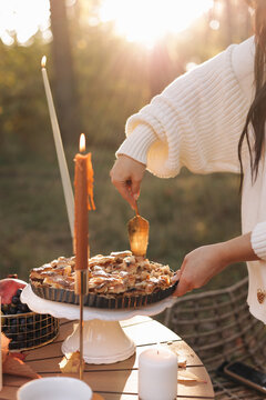 A woman in a cozy sweater serves a rustic apple pie on a pedestal during an autumn outdoor picnic, surrounded by candlelight, warm sunlight and seasonal fruit for a relaxed harvest mood.