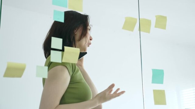 Business woman making a phone call while brainstorming and planning her project strategy, using colorful sticky notes on a glass board in a modern office