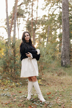 A young woman wearing a white dress and knee-high boots poses confidently in an autumn forest bathed in golden light, blending fashion and nature to evoke calm, style, and seasonal beauty.