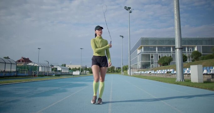 A young beautiful girl in sportswear and a cap with a skipping rope in her hands walks along the stadium's running track to train and exercise on a summer day