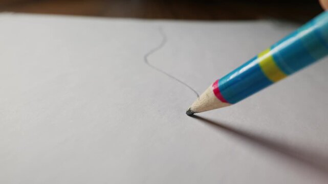 A hand draws a curved graphite line with a colorful striped pencil on a white sheet of paper. Concept of creativity, education, art, workspace writing, and childhood learning.