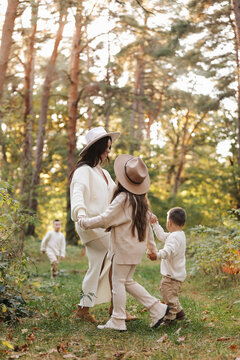 A mother and her children hold hands and play during a golden autumn walk through a sunlit forest, wearing cozy neutral sweaters and wide-brim hats, conveying warmth, connection and family 