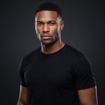 Serious young Black man in 20s looking at camera indoors against dark grey studio background wearing casual black t shirt with confident expression