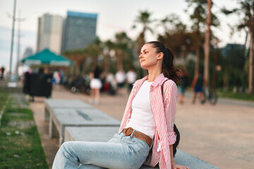 Fototapeta premium Young woman with long dark hair sits on a concrete bench wearing a white tee and striped shirt, relaxed in a sunny park with a distant city skyline.
