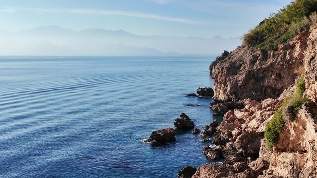 Turquoise mediterranean waves splash and swirl around jagged rocks beneath a rugged coastal cliff.