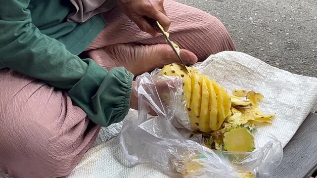 Close-up of skilled hands peeling a fresh pineapple with a traditional knife, creating a clean spiral pattern on the fruit. Tropical fruit preparation, craftsmanship, food processing.