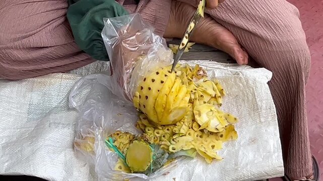 Close-up of skilled hands peeling a fresh pineapple with a traditional knife, creating a clean spiral pattern on the fruit. Tropical fruit preparation, craftsmanship, food processing.