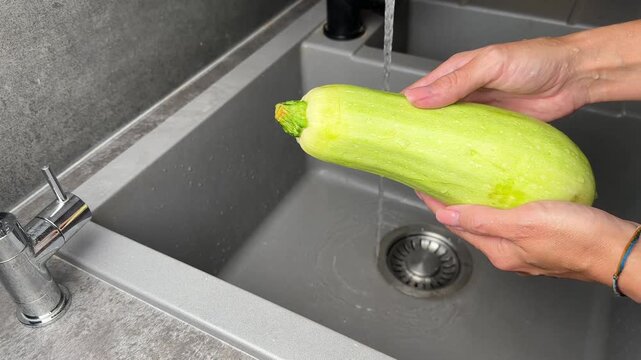 Video of a woman's hands washing a fresh green zucchini under running tap water in a grey composite kitchen sink, preparing healthy food, 4k