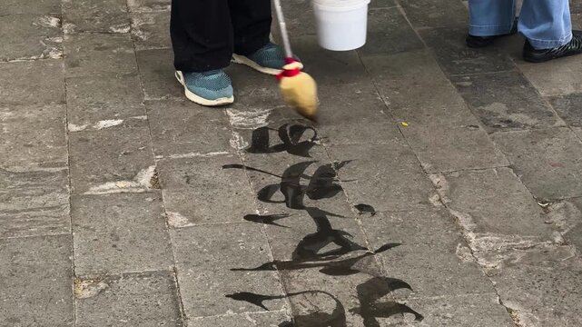 Person calligrapher practicing water calligraphy on stone pavement with large brush. Street artist writing Chinese characters with water. Traditional oriental art, mindful hobby and zen practice.