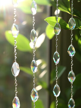 Crystal beads suncatcher hanging in front of a window with green leaves, sparkling in bright morning sunlight with rainbow reflections