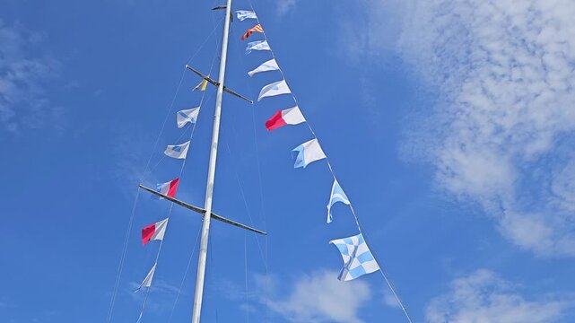 Nautical signal flags fluttering from a sailboat mast against a vivid blue sky with textured clouds, conveying a festive, summery maritime mood and the motion of wind on a clear day.