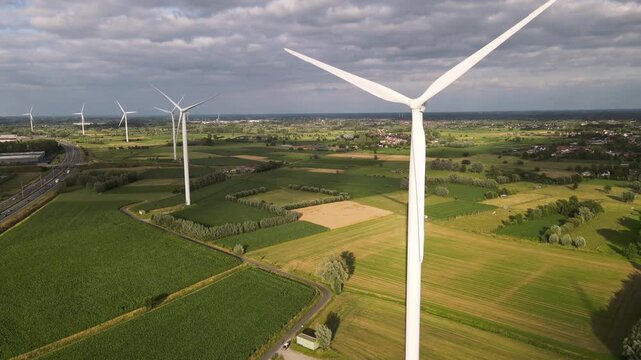 Aerial view of a wind turbine spinning in the foreground with more turbines across green patchwork fields and a highway, footage of renewable wind energy.
