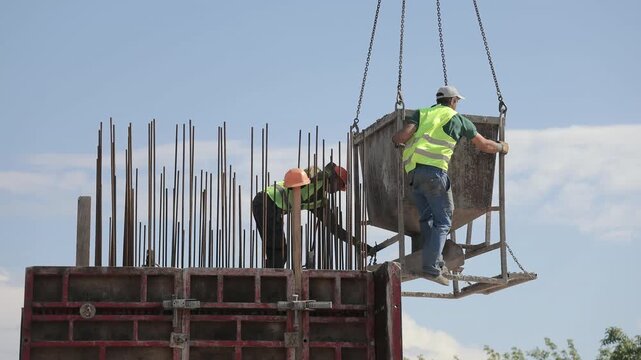 Construction workers guide a suspended concrete hopper above formwork, Crane chains and exposed reinforcing bars show active building operations