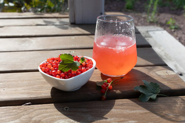 Close view of white bowl filled with glossy red currants topped with fresh mint leaf placed beside chilled pinkish red currant drink in glass on weathered wooden table outdoors, sunlit highlights on © olga_sova