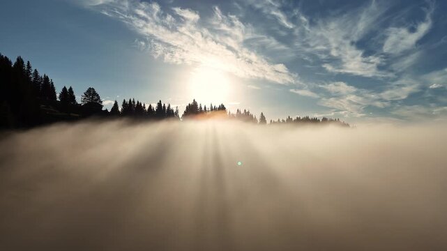 Bright sunrise breaks through dense fog above a dark mountain forest, Aerial view captures radiant sunbeams and wispy clouds across the blue sky