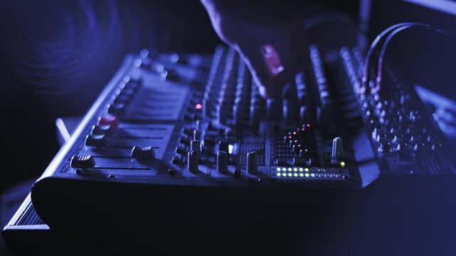 Low angle side view of a person's hand actively mixing audio on a console. Faders and knobs are adjusted as LED level meters fluctuate in a dark, blue-lit room or live venue.