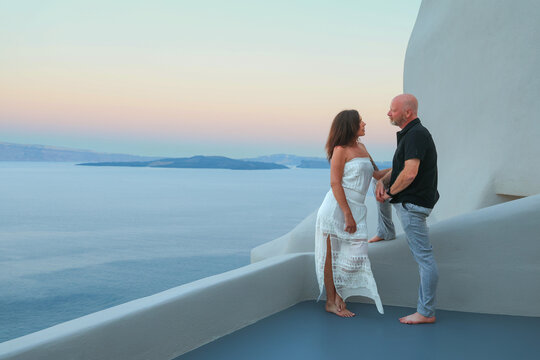 A couple stands on a white Santorini terrace at sunset, sharing a quiet moment above the Aegean Sea with soft pastel light and serene Mediterranean atmosphere in Greece.