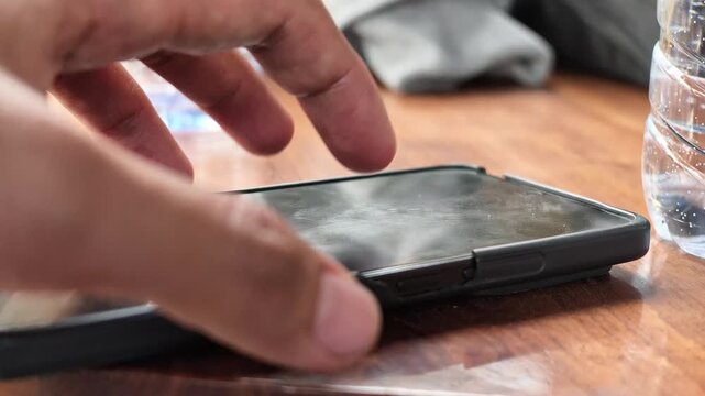 Slow motion detailed shot of a finger impatiently tapping on a black smartphone screen laying on a wooden table, reflecting communication delays or boredom.
