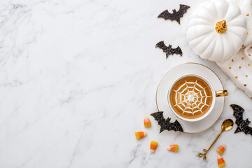 Halloween coffee cup with spider web latte art, white pumpkin, candy corn and bats on white marble background with copy space flat lay