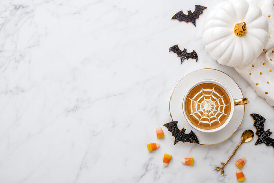 Halloween coffee cup with spider web latte art, white pumpkin, candy corn and bats on white marble background with copy space flat lay