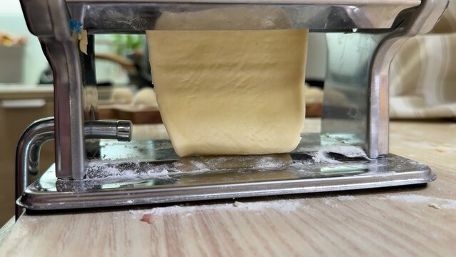 Close view of pasta machine rolling a sheet of dough on a kitchen counter, flour dusted surface and metal rollers suggest home cooking and preparation