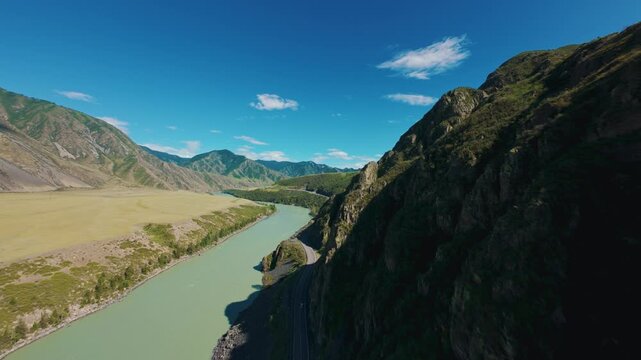 Aerial tracking shot of turquoise river winding through mountain valley beside cliff road with forward pan motion Chuysky tract Altai, Siberia, Russia.