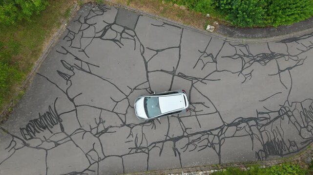 Silver car parked on an asphalt road riddled with cracks, highway patched with dark tar, street repairs, deterioration, dilapidated stretches of pavement