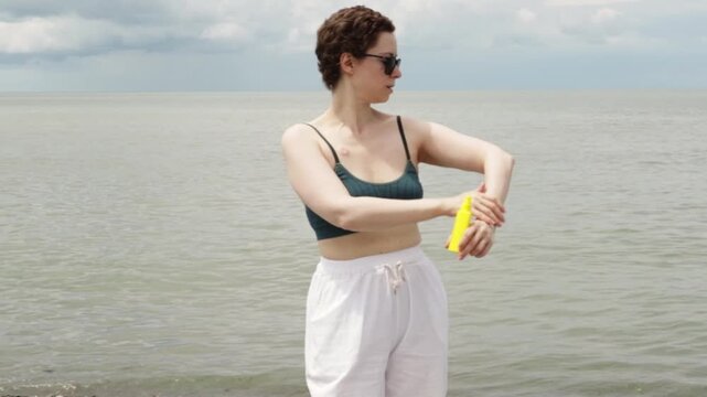 cancer survivor applies sunscreen to her shoulder at the seaside, demonstrating careful protection from sun exposure during summer recovery and post treatment wellness
